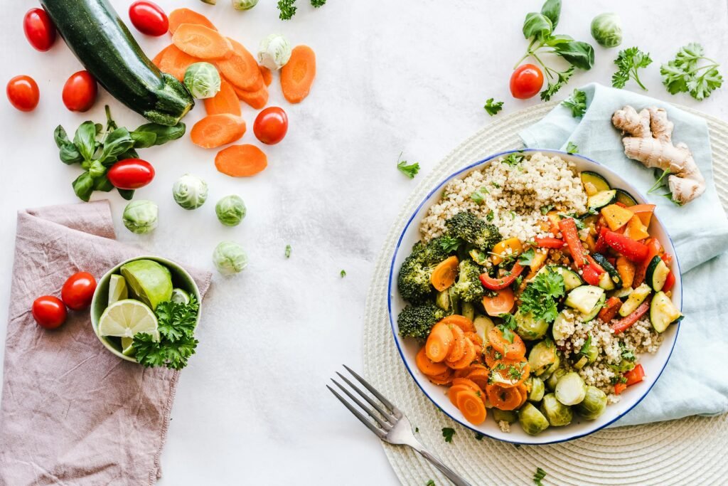 A healthy meal is presented on a plate. It's a quinoa salad with lots of vegetables like carrots, broccoli, and zucchini. Some vegetables are displayed on the table, along with a bowl of cut limes and fresh ginger.