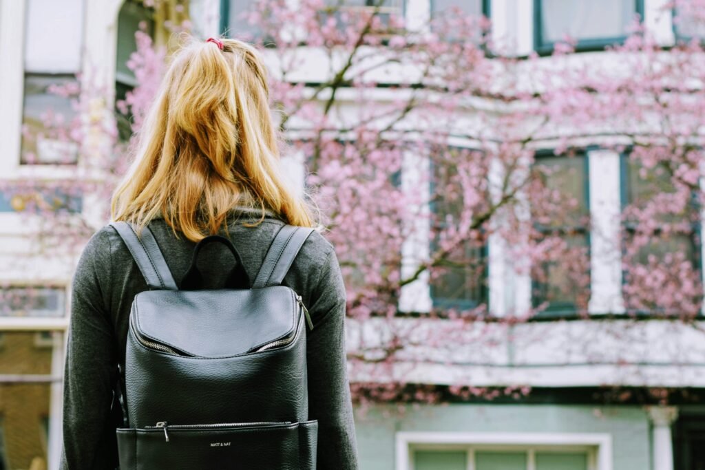 A young woman with a black leather backpack is standing in front of a building and a cherry tree full of flowers.