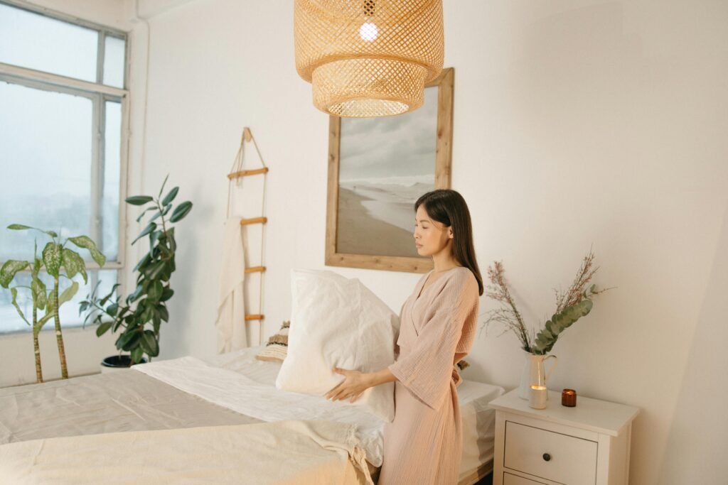 Asian woman arranging pillow in a cozy, minimalistic bedroom setting.
